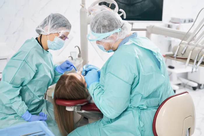 Doctors wearing protective face masks and safety medical suits during oral surgery for woman in dental clinic